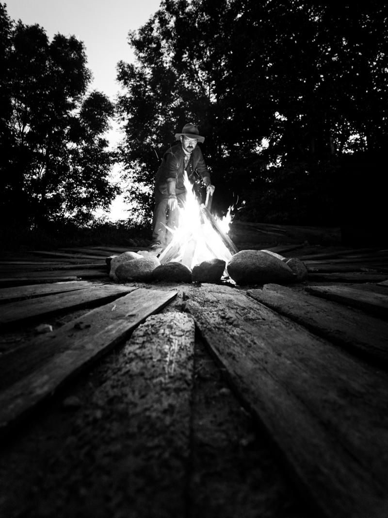 A man in a hat tending a campfire on a wooden platform at dusk