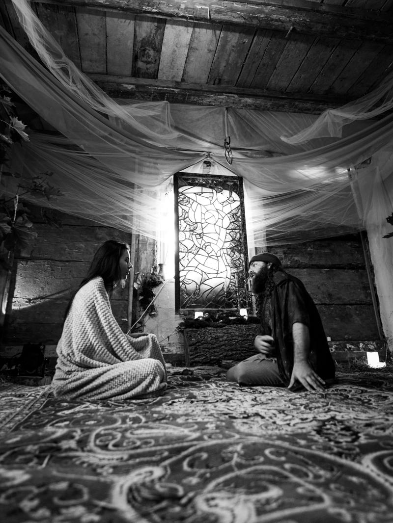 Two people sitting on rugs in deep conversation beneath draped fabric and a stained-glass window