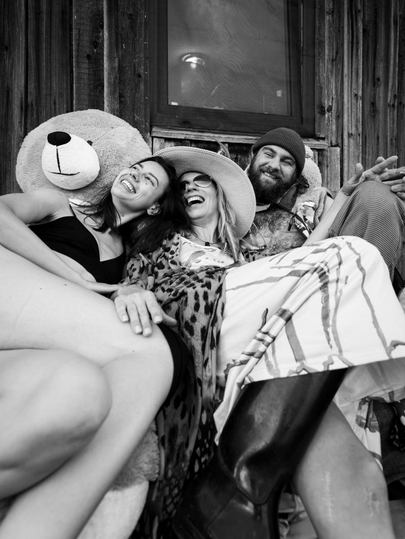 Three friends laughing together on a porch next to a giant teddy bear