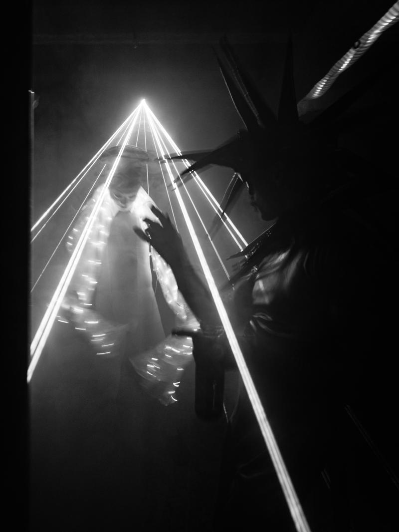 A costumed figure in a spiked headdress among beams of laser light and smoke